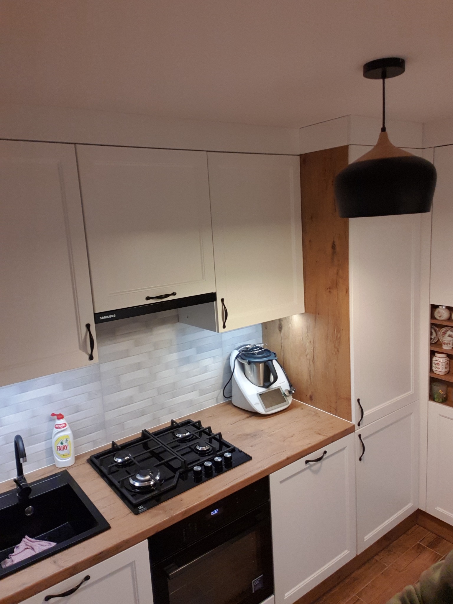 Biała kuchnia z drewnianym blatem, czarnym zlewem i płytą gazową, oświetlona wiszącą lampą; widoczny robot kuchenny i akcesoria.