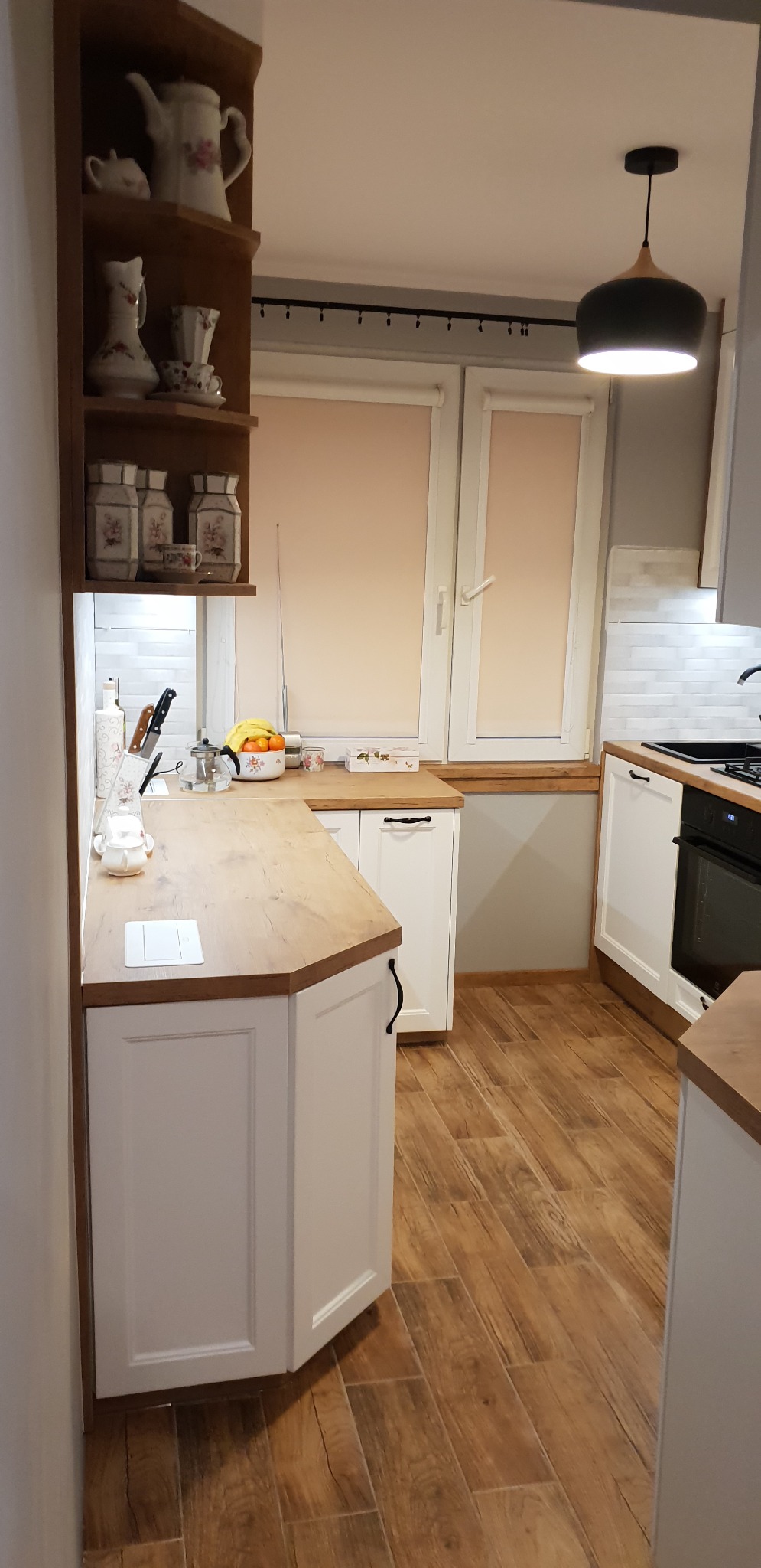 Jasna kuchnia z drewnianym blatem i podłogą imitującą drewno; na półkach dekoracyjne naczynia i pojemniki.