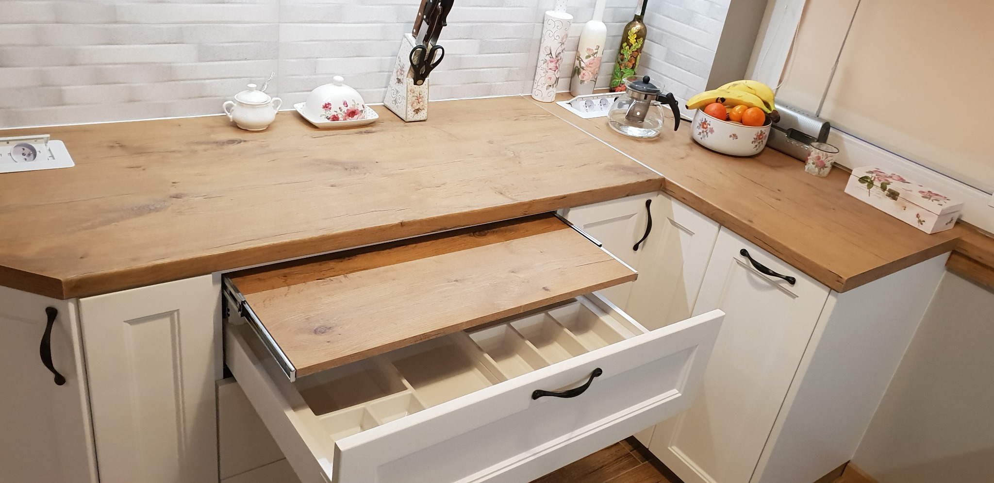 Wysuwana szuflada z przegródkami w białej szafce kuchennej z drewnianym blatem, widoczne akcesoria kuchenne i owoce.