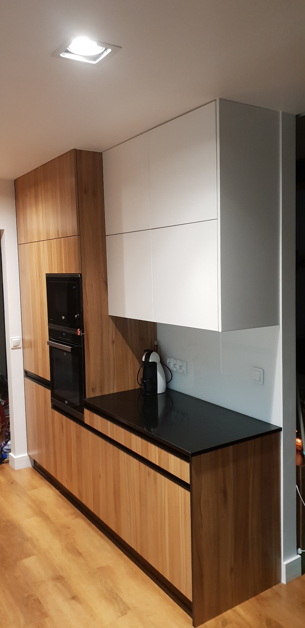 Nowoczesna kuchnia z drewnianymi szafkami dolnymi i białymi górnymi, czarny blat, wbudowany piekarnik i ekspres do kawy na blacie.