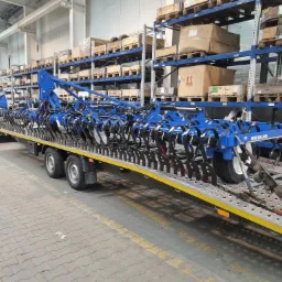 Niebieski agregat uprawowy New Holland zabezpieczony pasami na żółtej platformie lawety, widok z boku na tle magazynu z regałami.