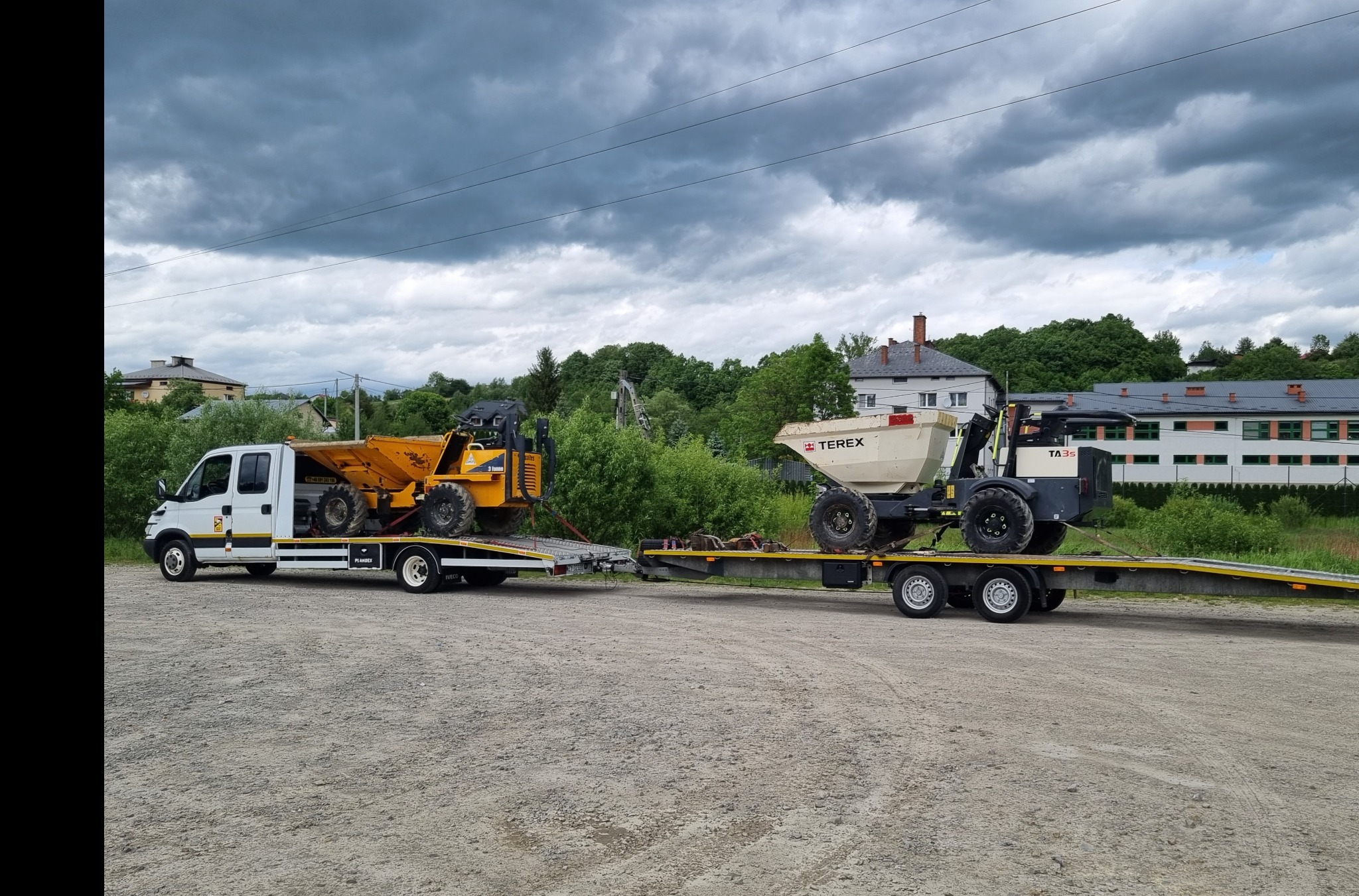 Laweta przewozi dwie mini wywrotki budowlane na tle pochmurnego nieba i budynków.