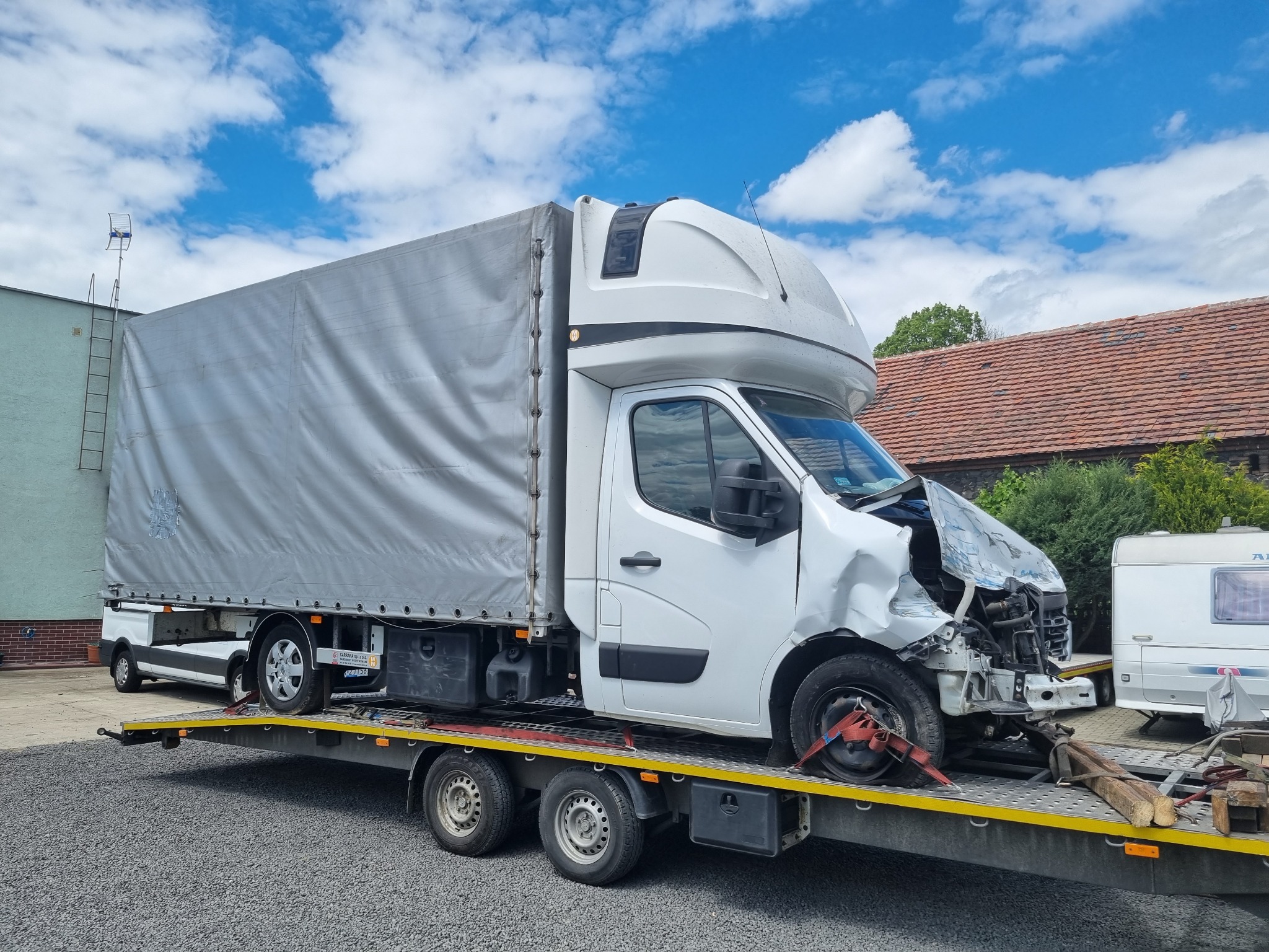 Uszkodzony biały bus z plandeką przewożony na lawecie, widoczne uszkodzenia przodu pojazdu, tło to budynek i błękitne niebo z chmurami.