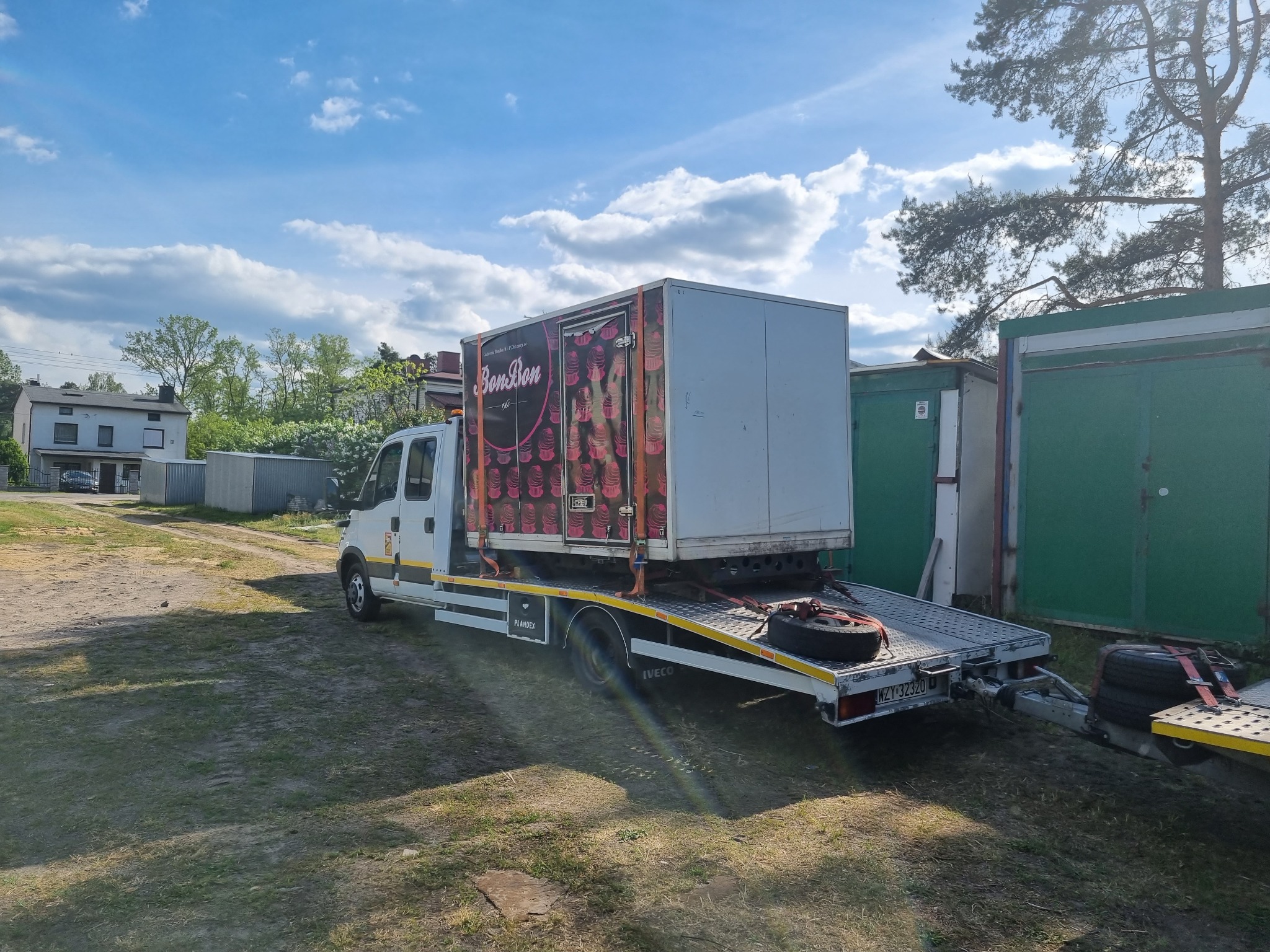 Laweta z platformą transportową przewozi białą kontenerową zabudowę z reklamą BonBon na tle budynków i zieleni.