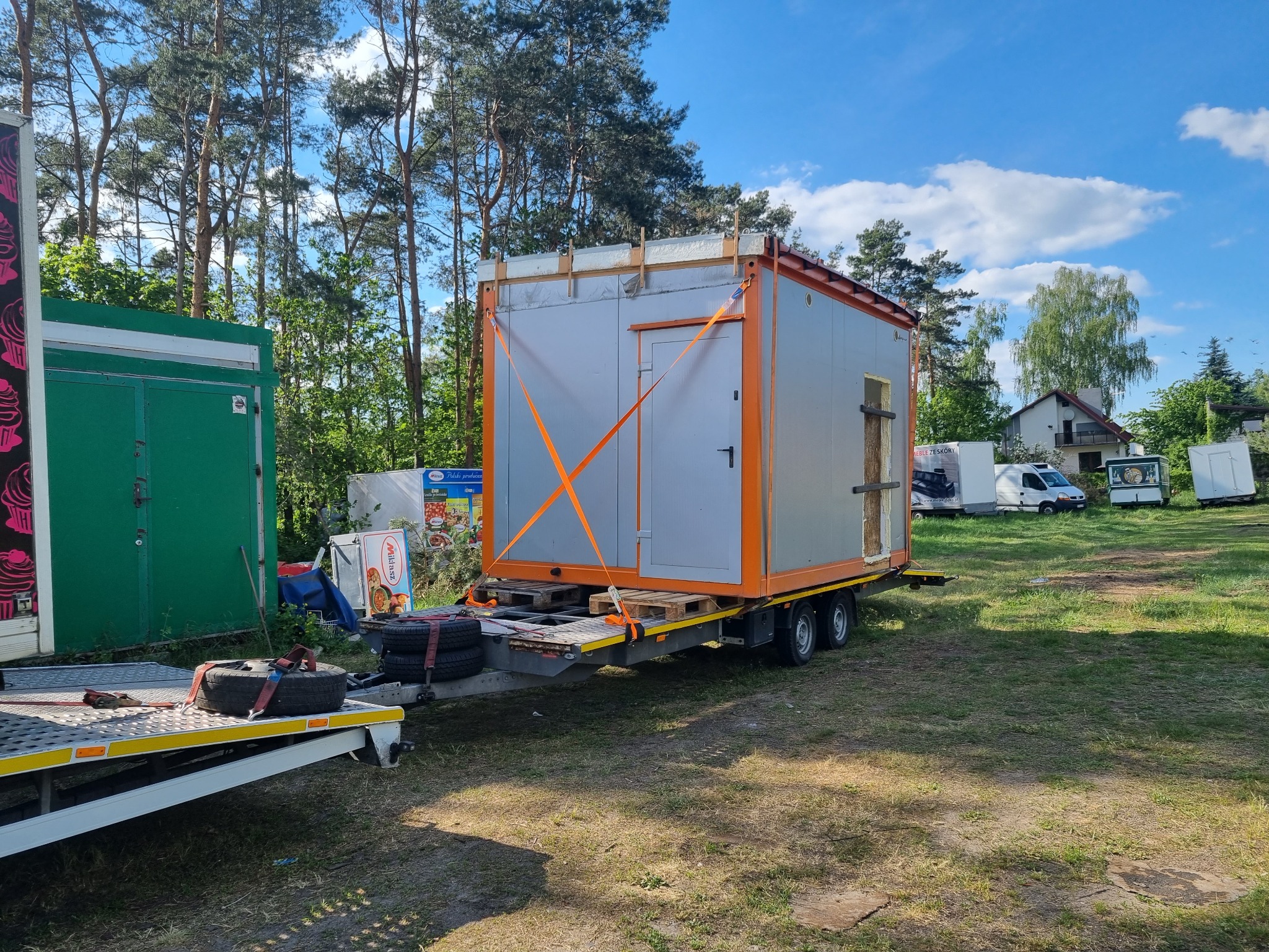 Szary kontener z pomarańczowymi obramowaniami, zabezpieczony pomarańczowymi pasami transportowymi, umieszczony na przyczepie lawety w plenerze.