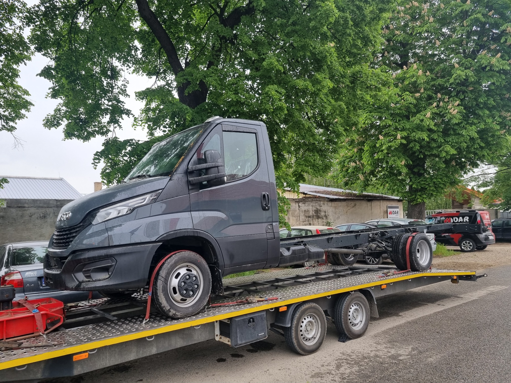 Szara kabina samochodu dostawczego bez zabudowy, zabezpieczona czerwonymi pasami, transportowana na żółtej lawecie w pochmurny dzień, z zielonymi drzewami w tle.