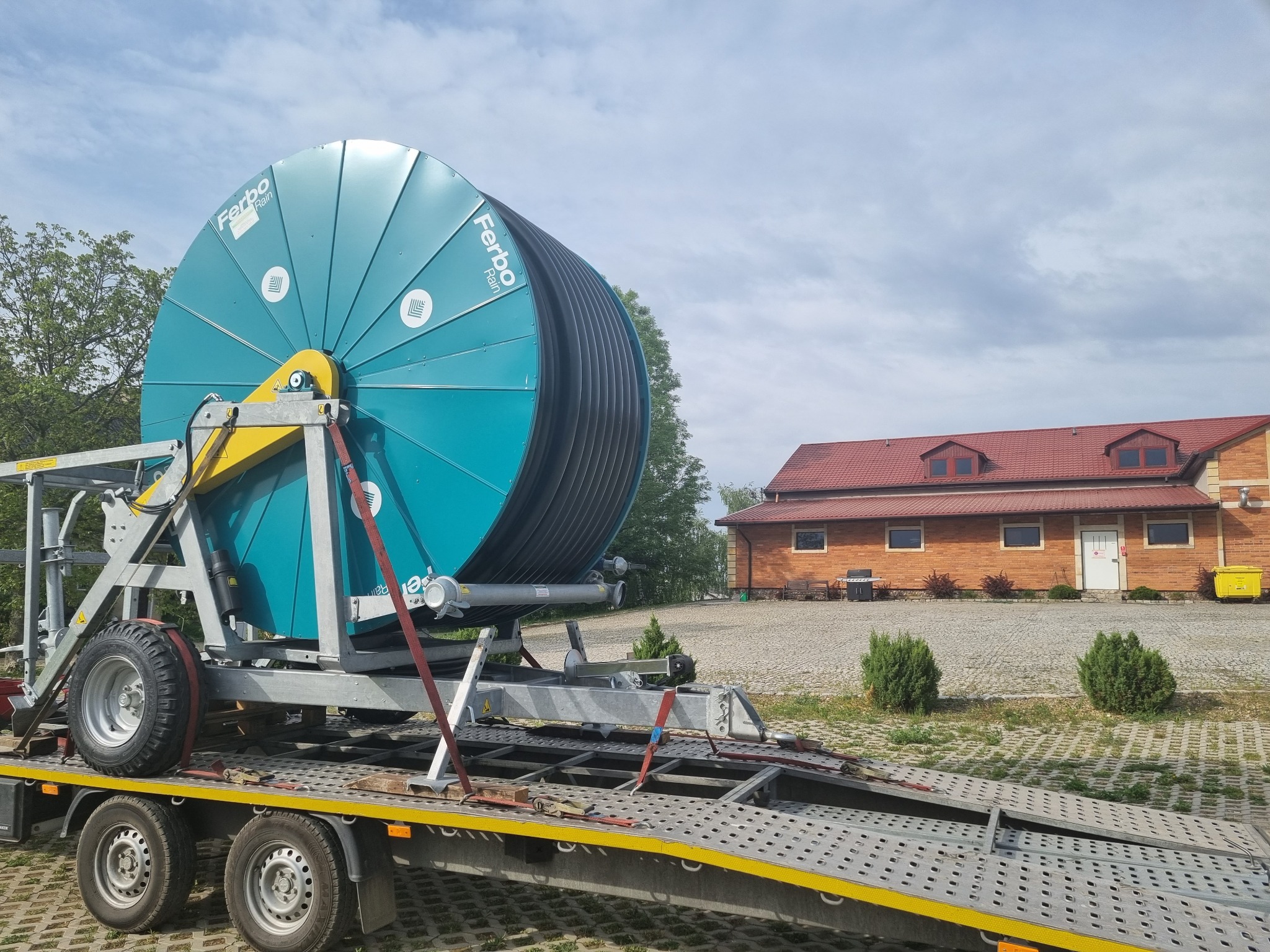 Niebieska szpula nawadniająca Ferbo Rain na platformie lawety z widoczną konstrukcją stalową i kołami, na tle budynku z czerwonej cegły i brukowanej nawierzchni.