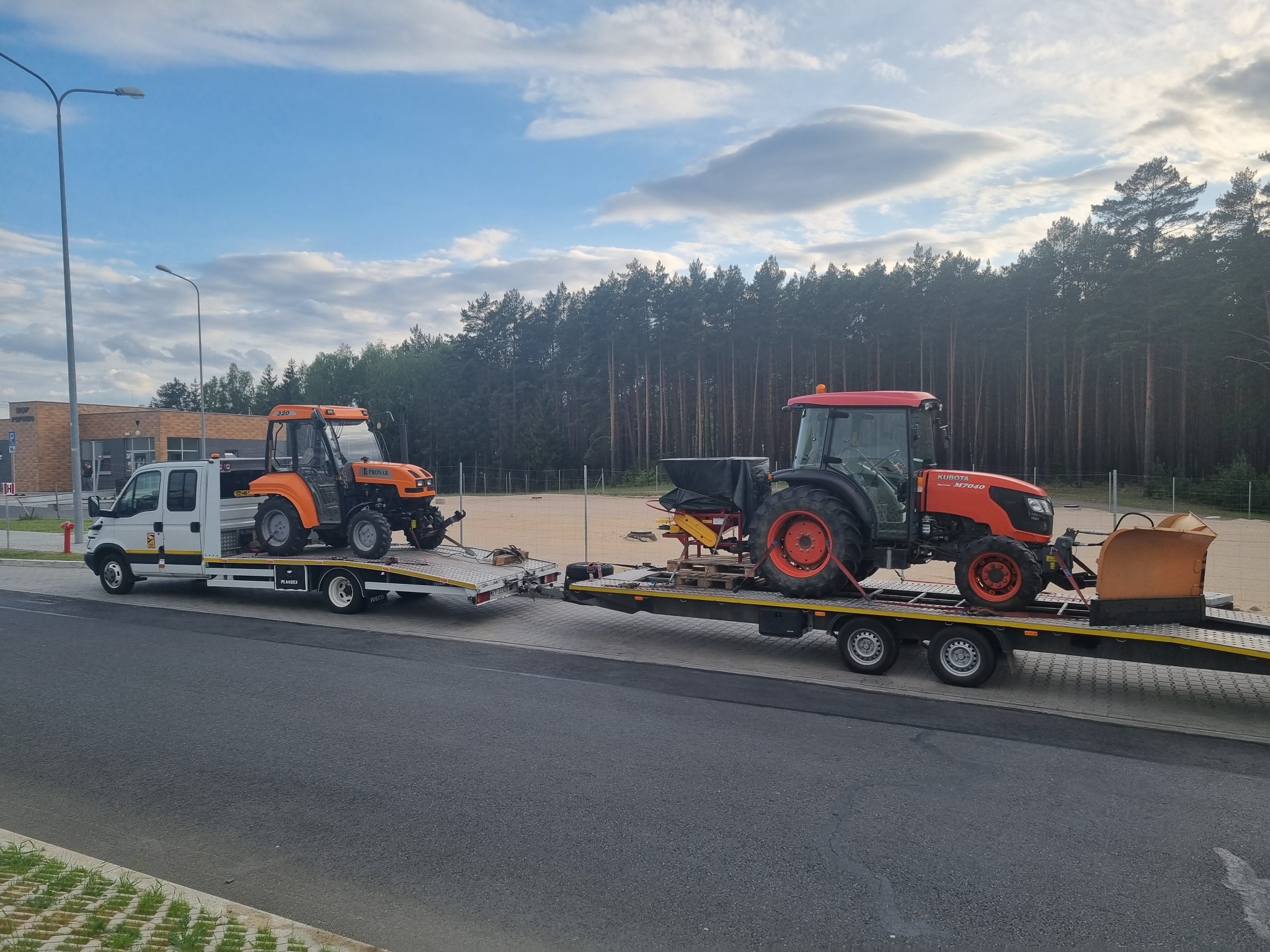 Dwa traktory, jeden pomarańczowy Pronar 320 i drugi czerwony Kubota, przewożone na lawetach ciągniętych przez białą ciężarówkę Iveco, na tle drzew i budynku.