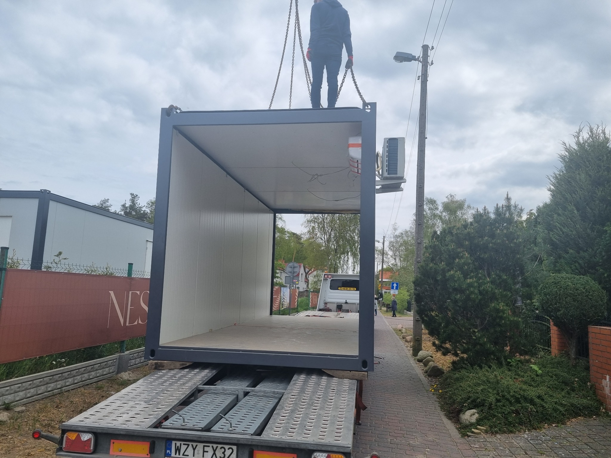 Kontener modułowy z klimatyzatorem transportowany na lawecie, podnoszony przez dźwig, widok z tyłu lawety na tle zabudowy miejskiej.