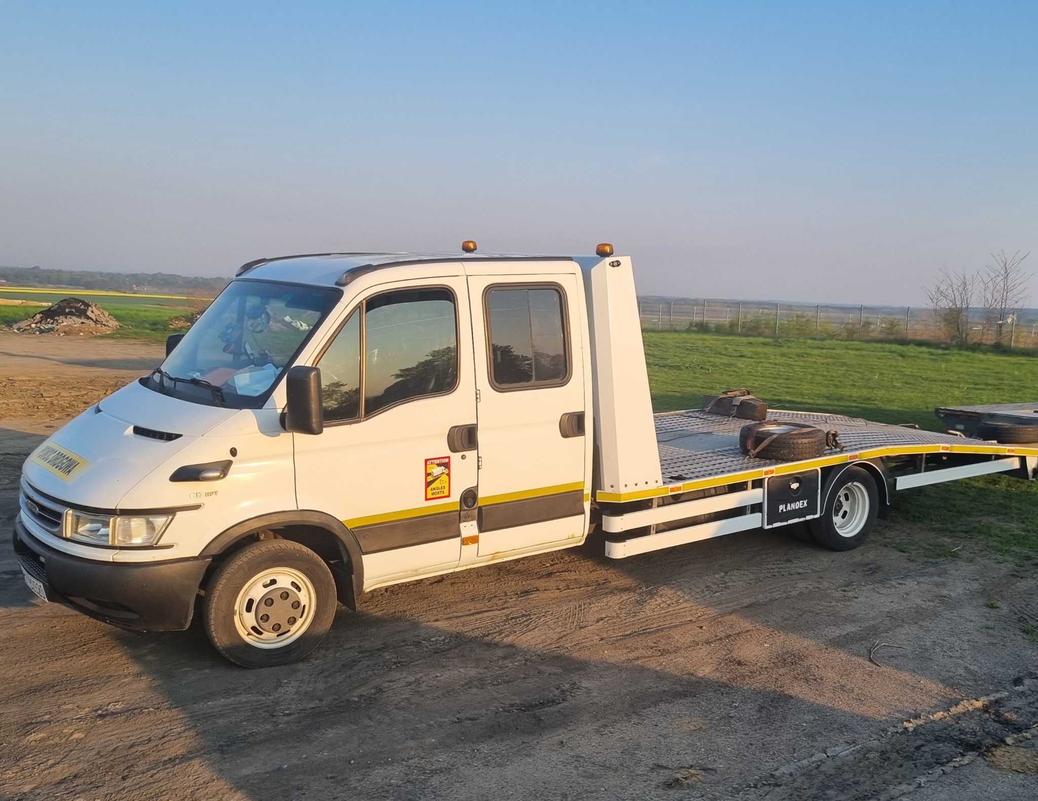 Biała laweta Iveco z żółtymi elementami ostrzegawczymi, zaparkowana na ziemistym placu z widokiem na pola w oddali.