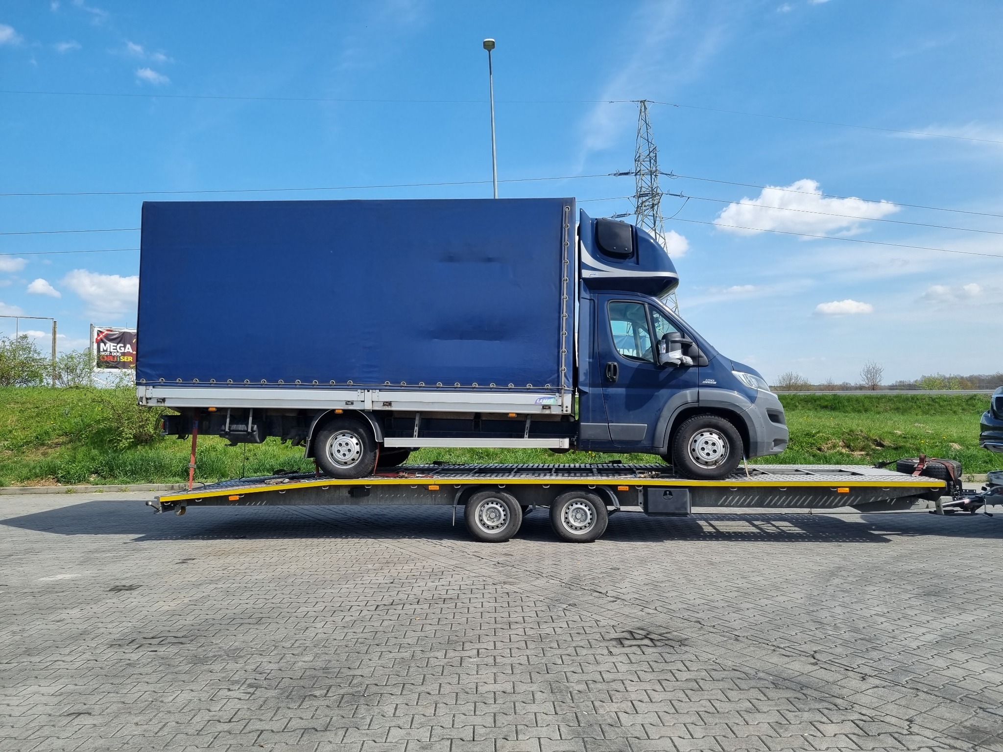 Niebieski bus z plandeką przewożony na żółtej lawecie na tle błękitnego nieba z chmurami i słupów wysokiego napięcia.