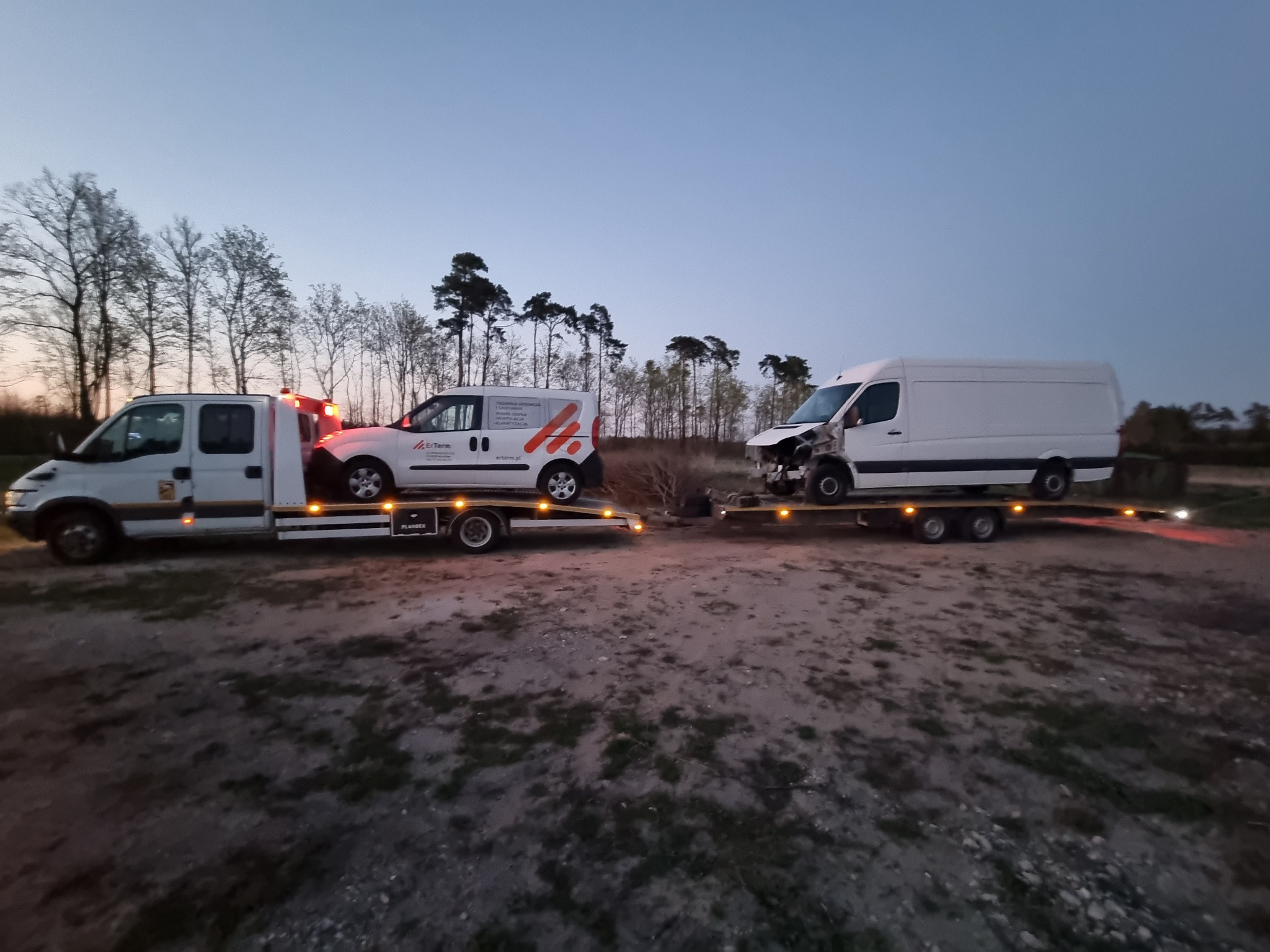 Dwa samochody lawetowe przewożące uszkodzone busy na tle leśnego krajobrazu o zmierzchu.