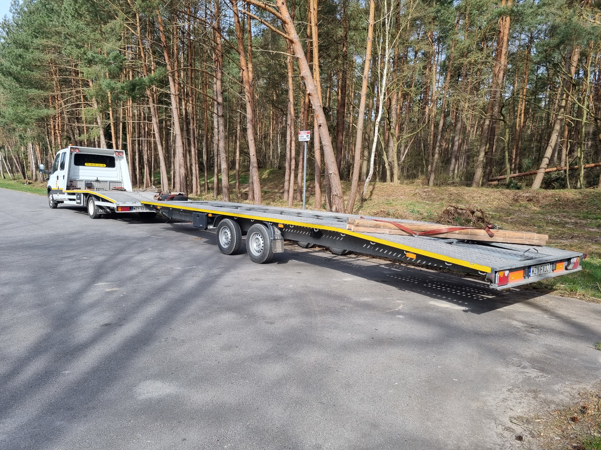 Biała laweta z drewnianymi belkami na platformie, zaparkowana na asfaltowej drodze w pobliżu lasu sosnowego.