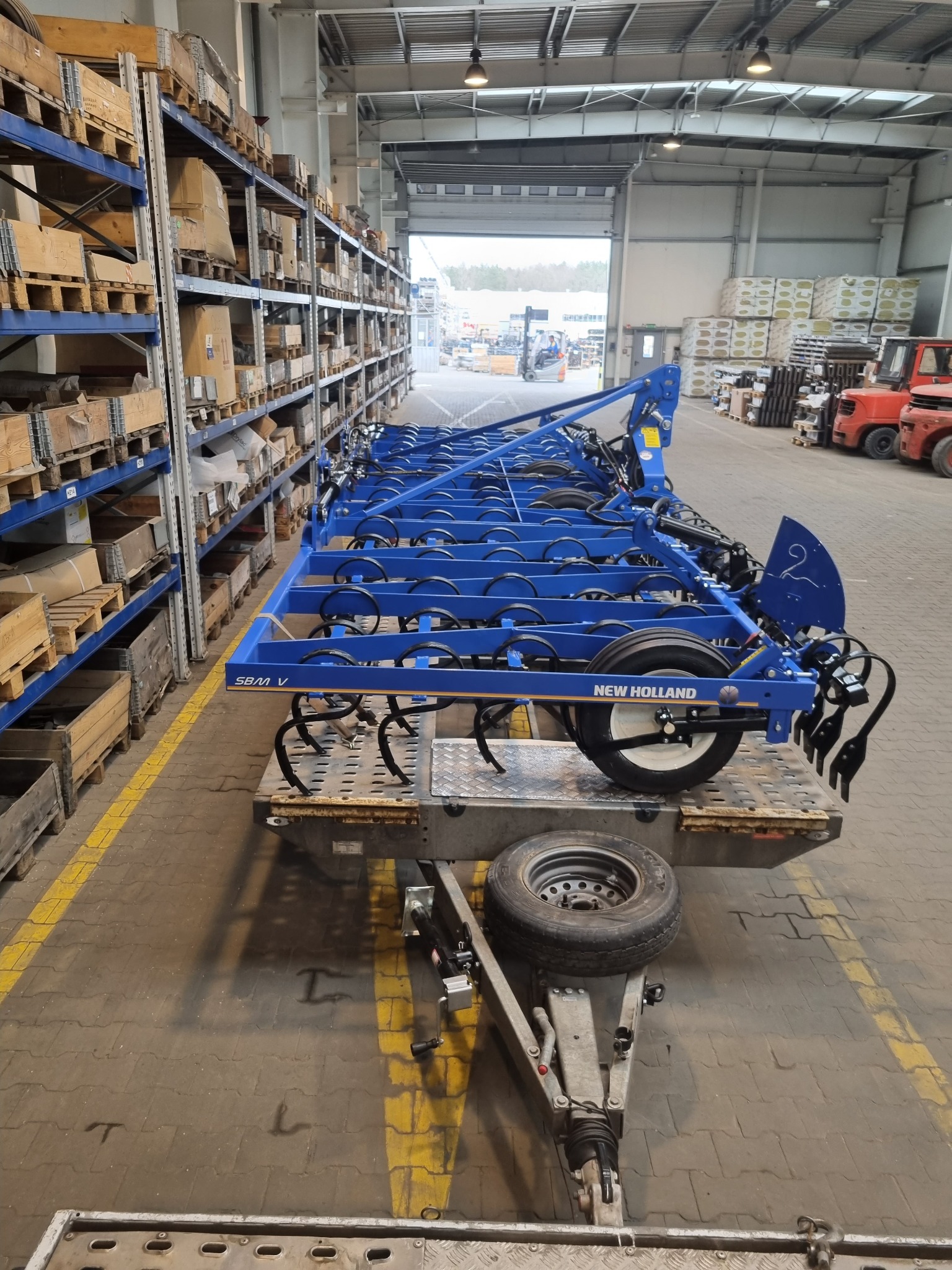 Niebieski agregat uprawowy New Holland SBM V na niskopodwoziowej platformie transportowej z zapasowym kołem, w hali magazynowej z regałami i wózkiem widłowym w tle.