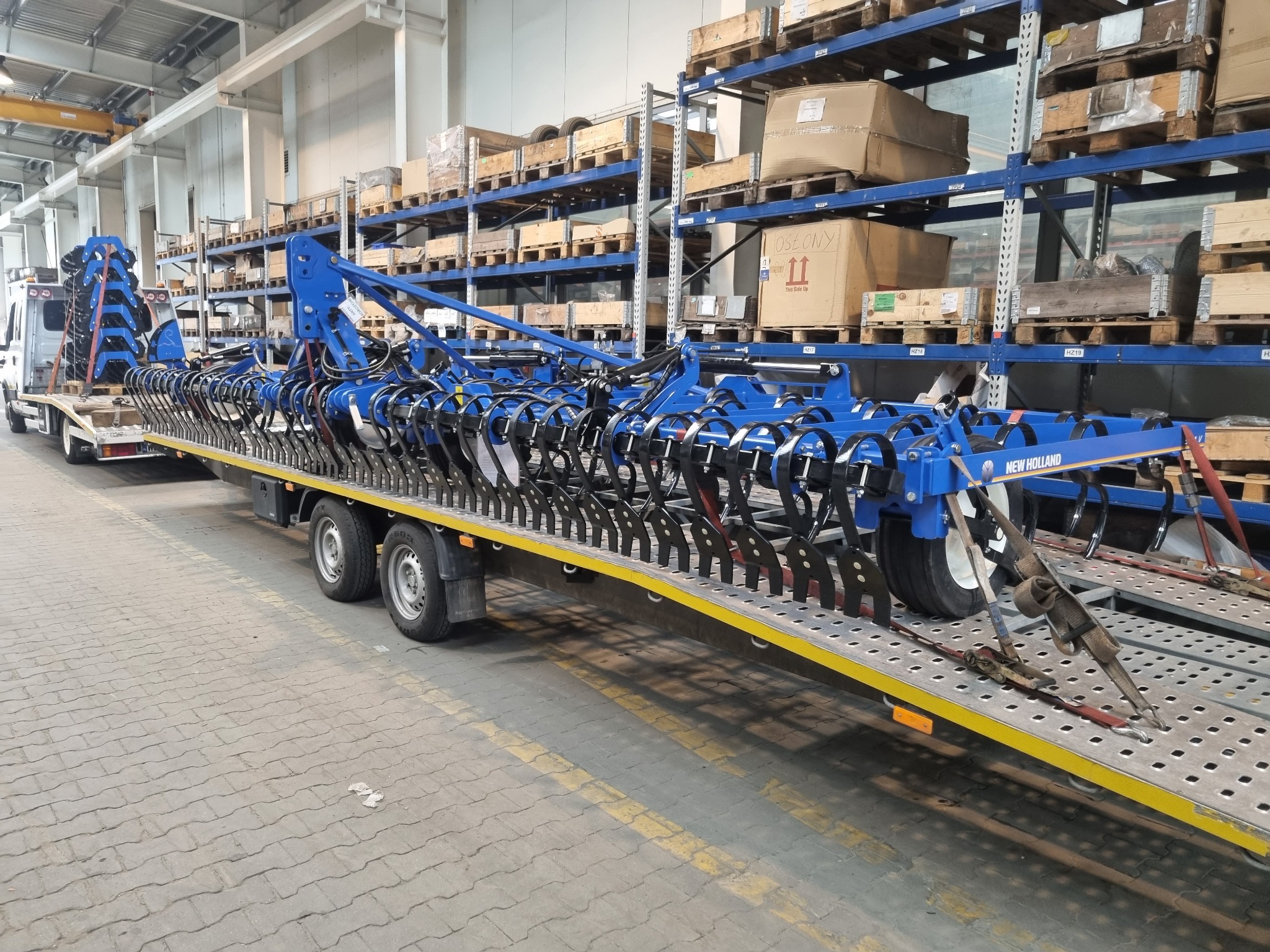 Niebieski agregat uprawowy New Holland zabezpieczony pasami na żółtej platformie lawety, widok z boku na tle magazynu z regałami.