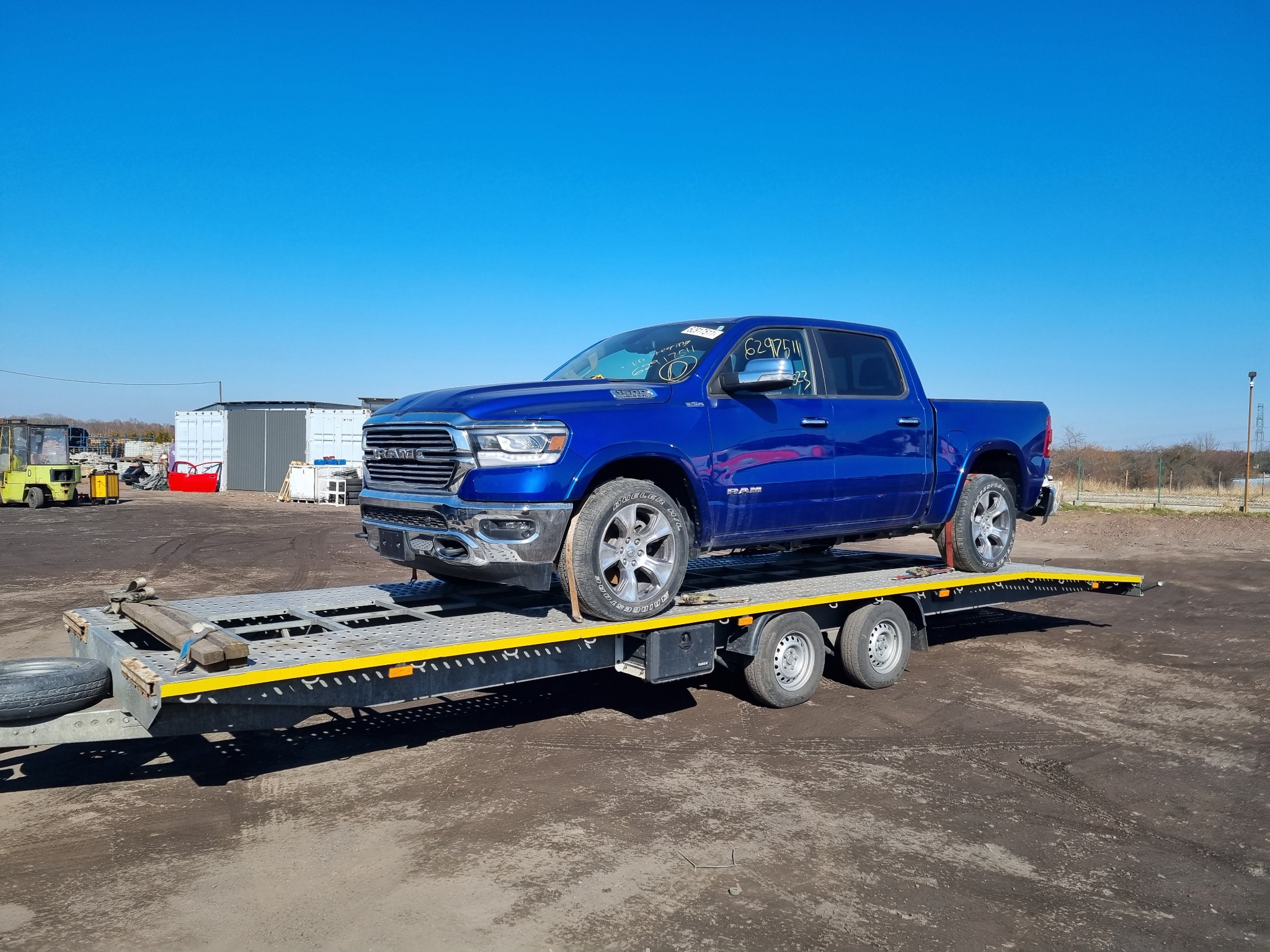 Niebieski pickup RAM na ażurowej lawecie z żółtymi elementami zabezpieczającymi, widoczne napisy na przedniej szybie, tło stanowią budynki gospodarcze i błękitne niebo.