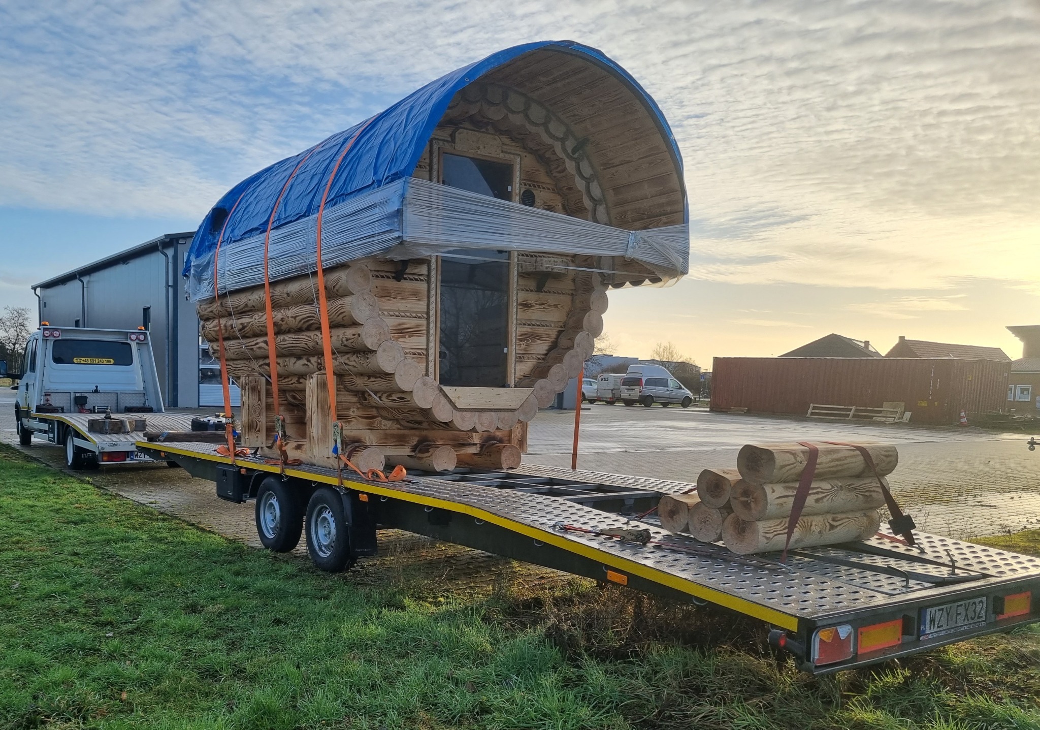 Laweta przewozi drewniany domek letniskowy z bali, zabezpieczony folią i plandeką, dodatkowo na lawecie leżą drewniane bale.