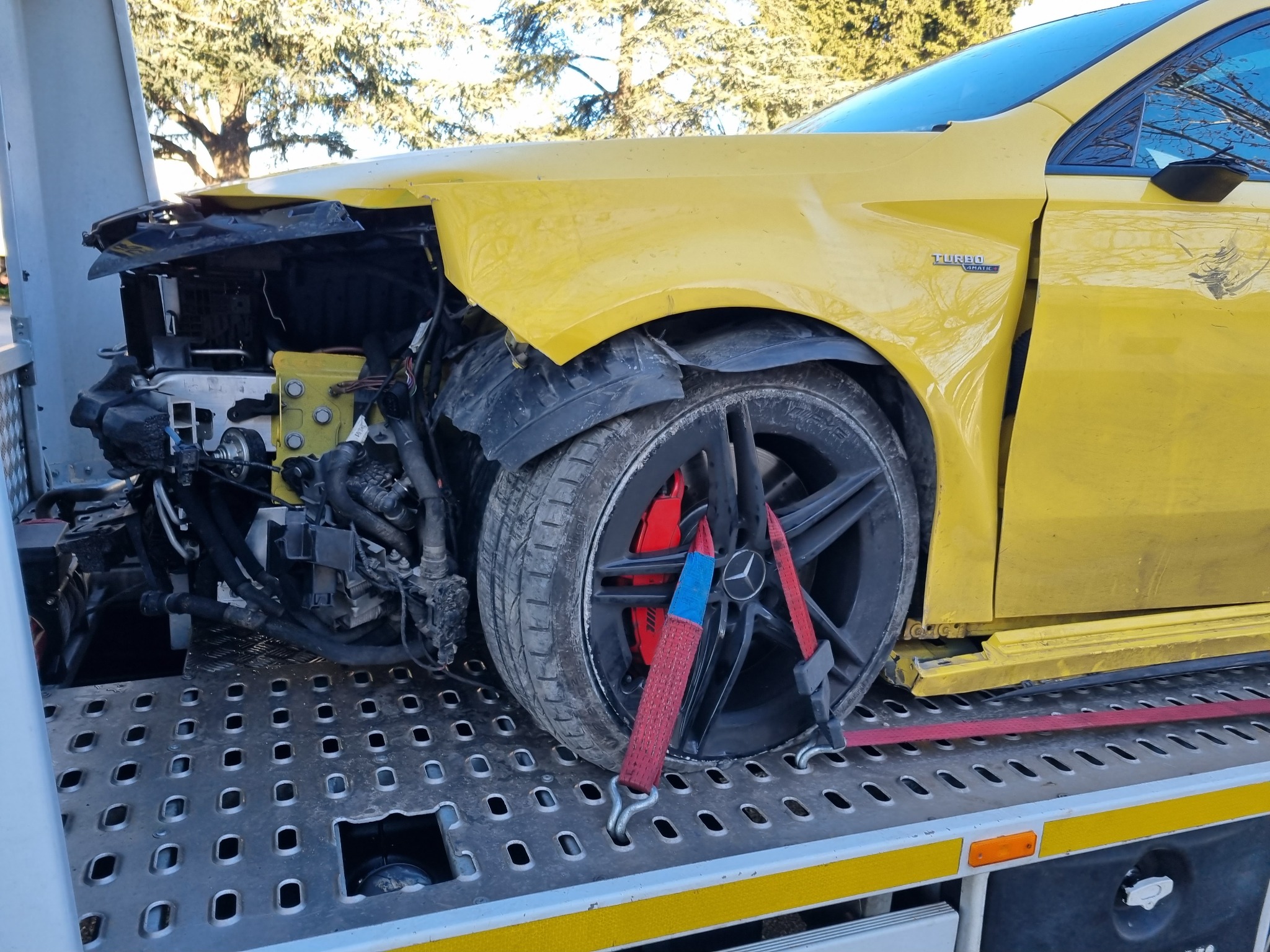 Żółty samochód z uszkodzonym przodem, zabezpieczony pasami transportowymi na platformie lawety. Widoczne elementy silnika i zawieszenia.