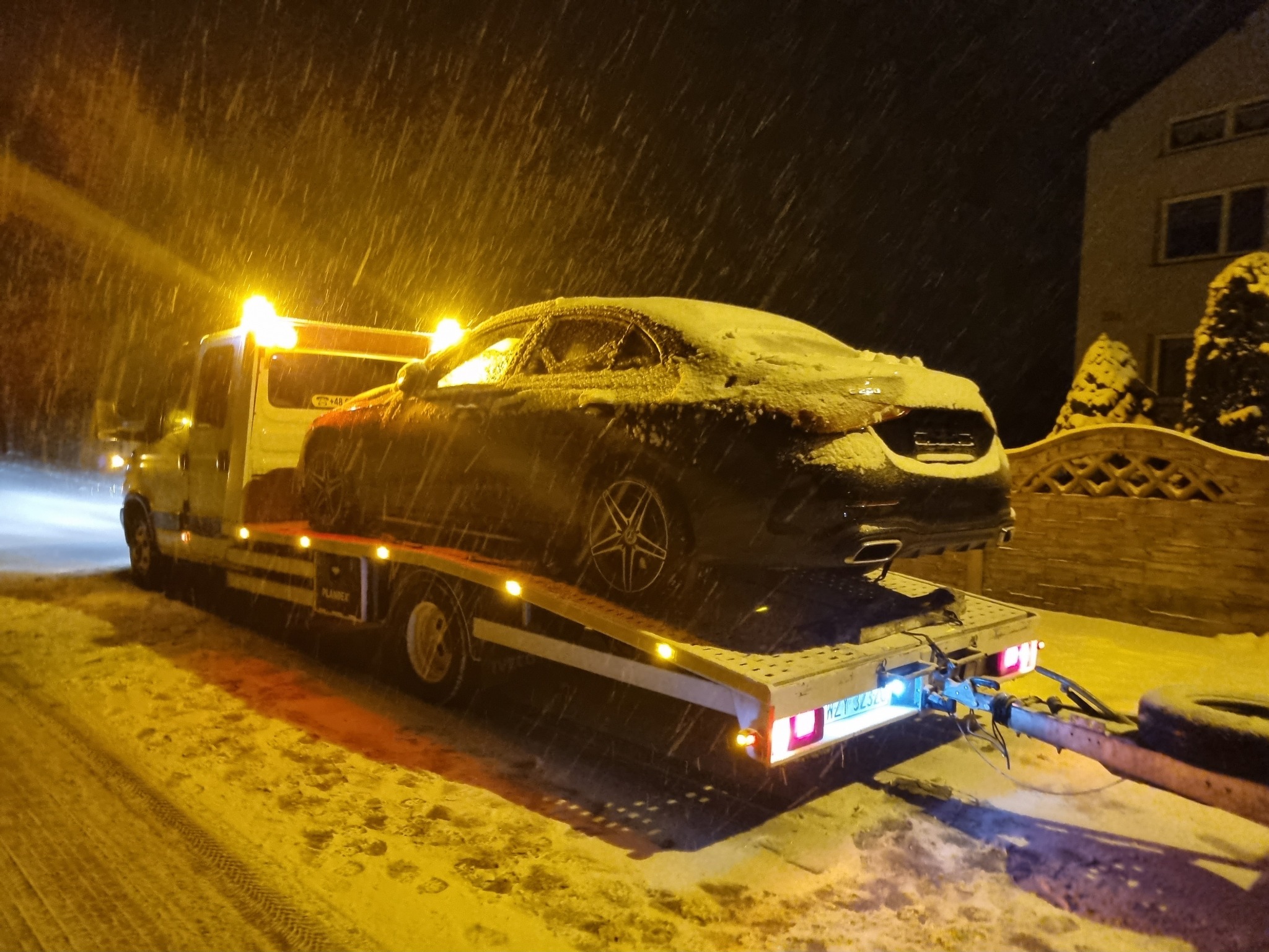 Czarny samochód osobowy pokryty śniegiem przewożony na lawecie w zimową noc podczas opadów śniegu, w tle oświetlony budynek.