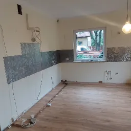 Wnętrze remontowanej kuchni z częściowo położoną mozaiką heksagonalną na ścianach, świeżo ułożonymi panelami podłogowymi i wystającymi rurami instalacji.