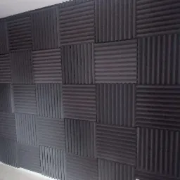 Ściana pokryta panelami akustycznymi w kolorze grafitowym, obok białe drzwi z szybkami na tle różowej ściany, widoczny fragment lampy i rośliny.