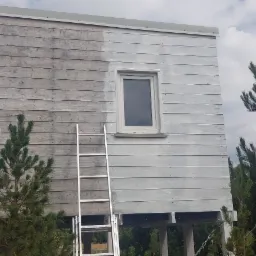 Drewniany domek na podwyższeniu w trakcie malowania; połowa ściany pomalowana na szaro, druga w surowym drewnie; aluminiowa drabina oparta o ścianę.