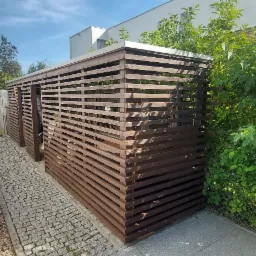 Drewniana konstrukcja osłaniająca śmietniki, wykonana z poziomych listew, na posesji z kostką brukową i bujną zielenią.