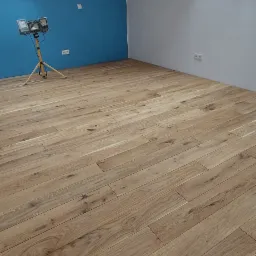 Jasne panele podłogowe ułożone w pokoju na poddaszu z niebieską ścianą i oknami dachowymi, oświetlone lampą budowlaną.