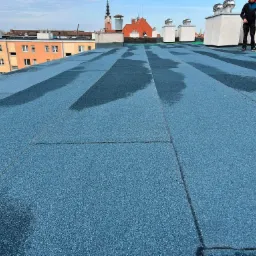 Nowe, niebieskie pokrycie dachu płaskiego z widocznymi łączeniami i plamami wody; w tle zabudowa miejska i dwaj pracownicy w trakcie inspekcji.