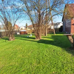 Rozległy trawnik z kilkoma drzewami, w tle zabudowa jednorodzinna i błękitne niebo. Na trawniku widoczne dwie osoby w białych kombinezonach.