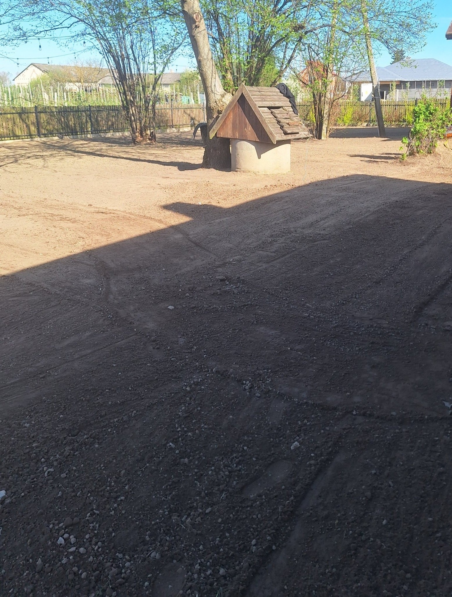 Obszerny, piaszczysty ogród z widoczną studnią z drewnianym daszkiem, otoczony drzewami i ogrodzeniem. Ciemny cień przecina dolną część kadru. Słoneczny dzień.