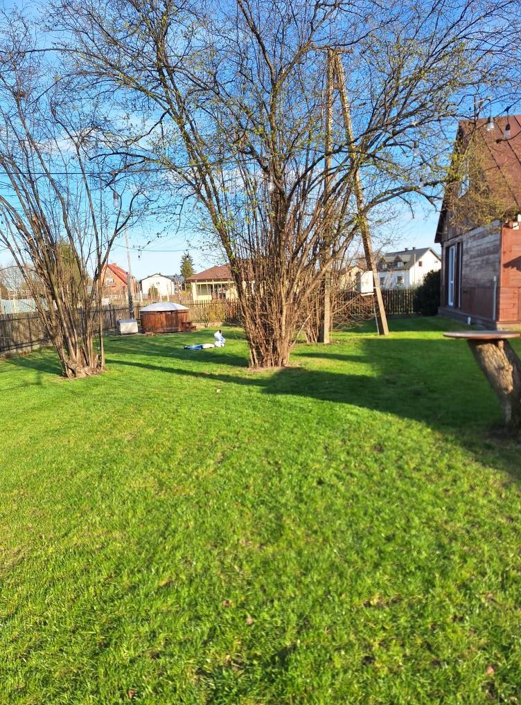 Rozległy trawnik z kilkoma drzewami, w tle zabudowa jednorodzinna i błękitne niebo. Na trawniku widoczne dwie osoby w białych kombinezonach.