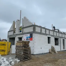 Wznoszenie ścian domu z bloczków Ytong, widoczne palety z materiałem, pracownicy na budowie, stan surowy otwarty, pochmurne niebo.