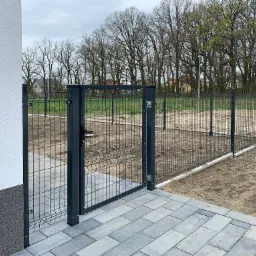 Nowoczesne ogrodzenie panelowe w kolorze grafitowym z furtką, zamontowane na posesji przy budynku z jasną elewacją i szarą kostką brukową.