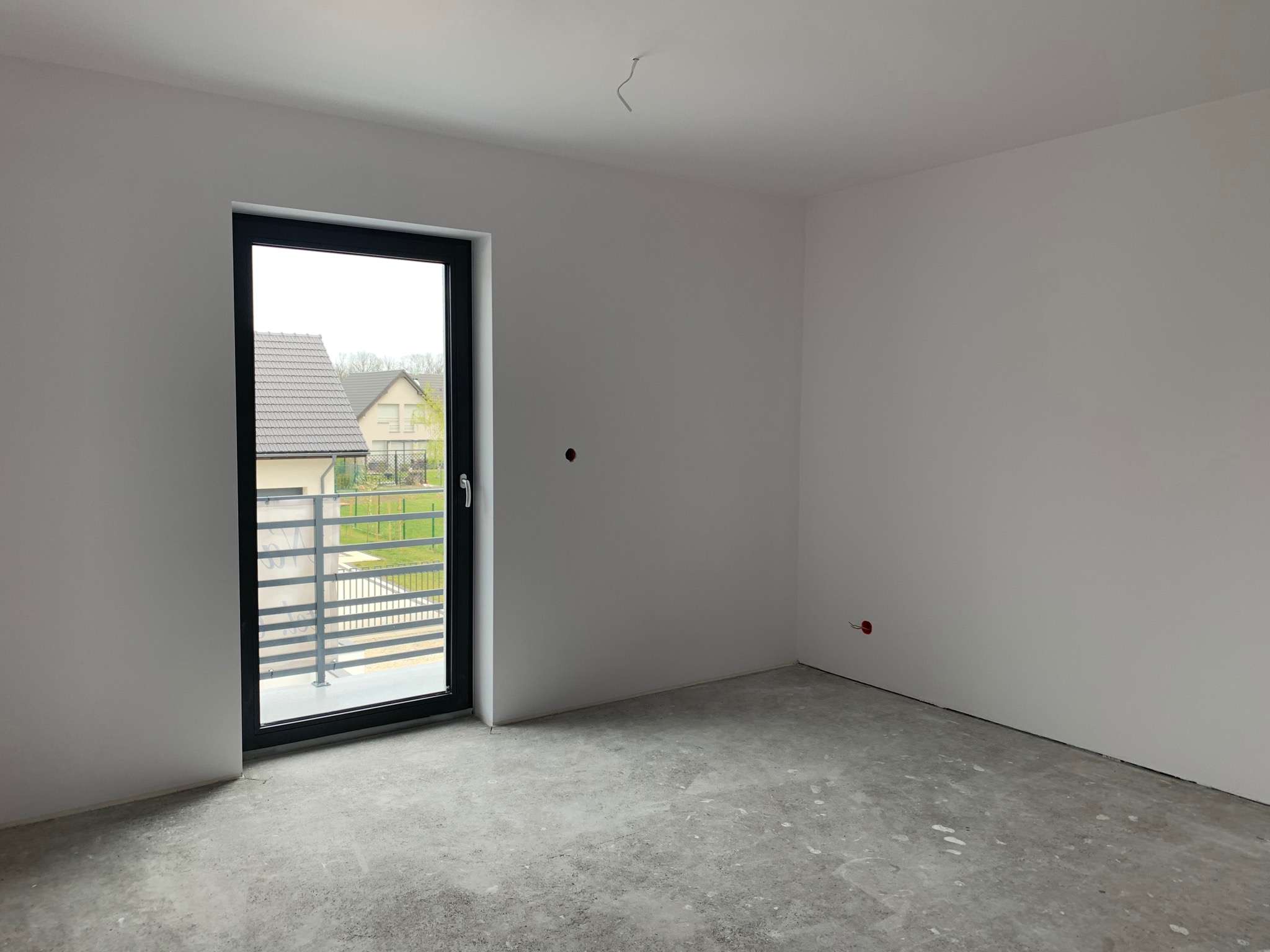 Wnętrze w stanie deweloperskim: surowe ściany pomalowane na biało, betonowa posadzka, czarne okno balkonowe z widokiem na ogród i domy sąsiadów, wystający kabel ze ściany.