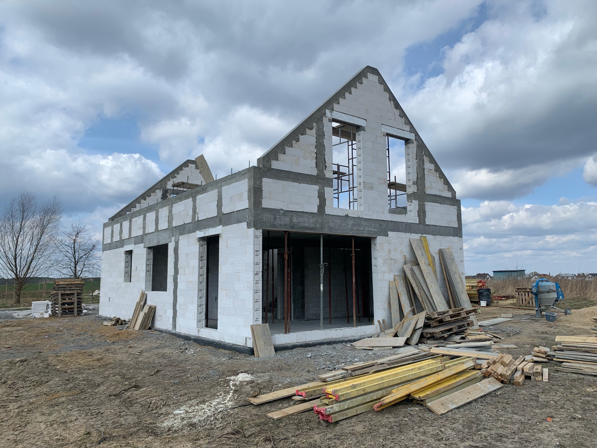 Surowy stan domu z białych bloczków, widoczne otwory okienne i drzwiowe, szalunki, drewniane podpory i materiały budowlane na placu budowy pod pochmurnym niebem.