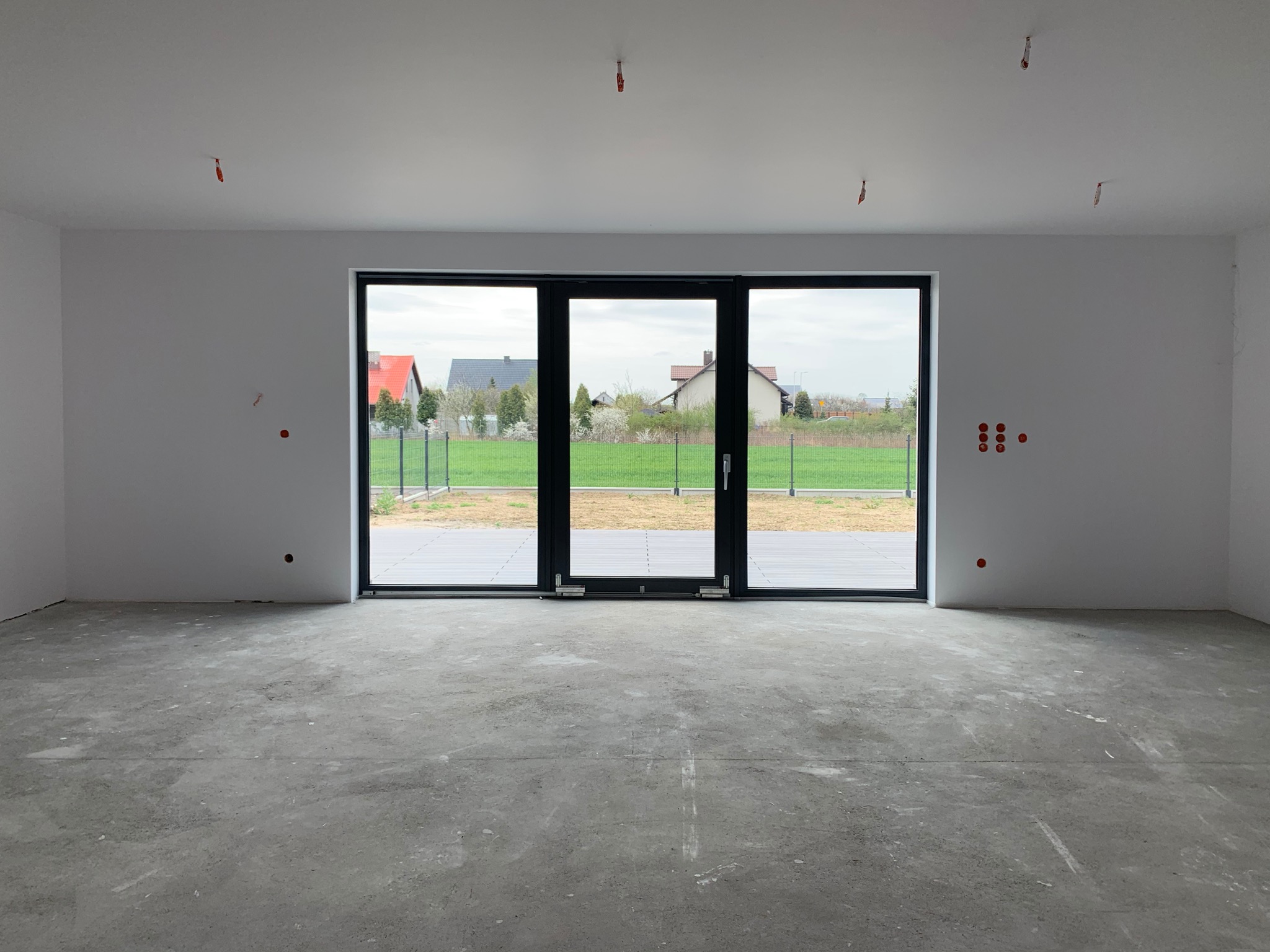 Surowe wnętrze domu w trakcie budowy z dużym oknem tarasowym, widoczne instalacje elektryczne na ścianach i suficie, betonowa wylewka podłogi oraz widok na ogród z domami w tle.