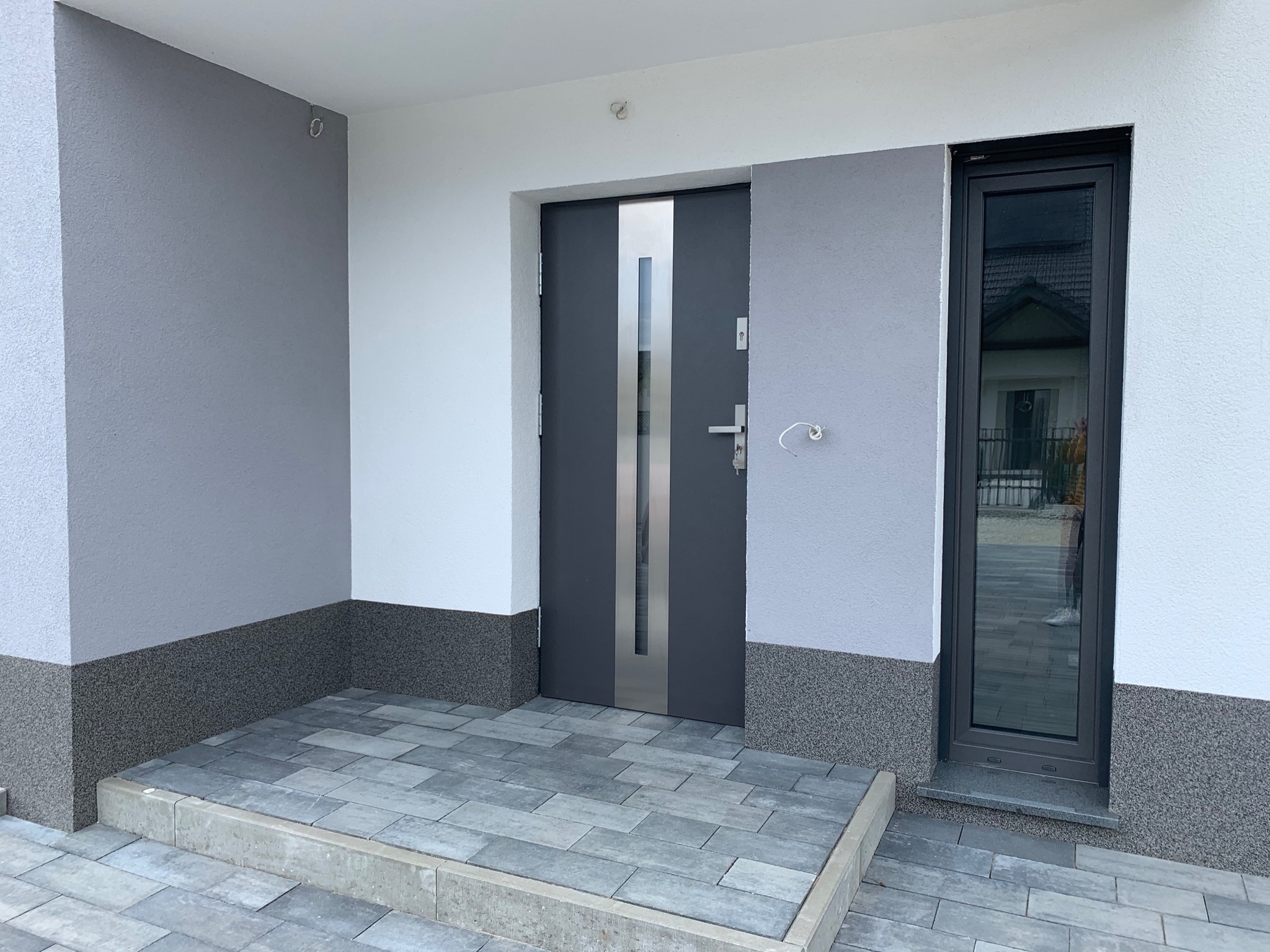Nowoczesne wejście do domu z szarymi ścianami o nierównej fakturze, ciemnoszarymi drzwiami z metalową wstawką i oknem z ciemną ramą, wyłożone szarą kostką brukową.