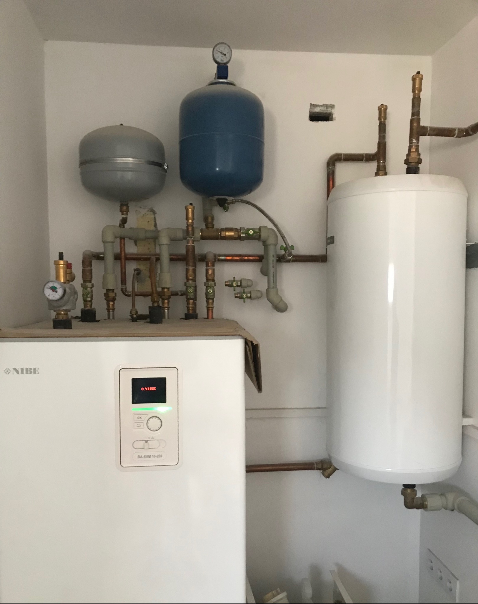 Biały piec grzewczy Nibe z szarym i niebieskim zbiornikiem wyrównawczym oraz białym zasobnikiem ciepłej wody, połączone miedzianymi rurami w kotłowni.
