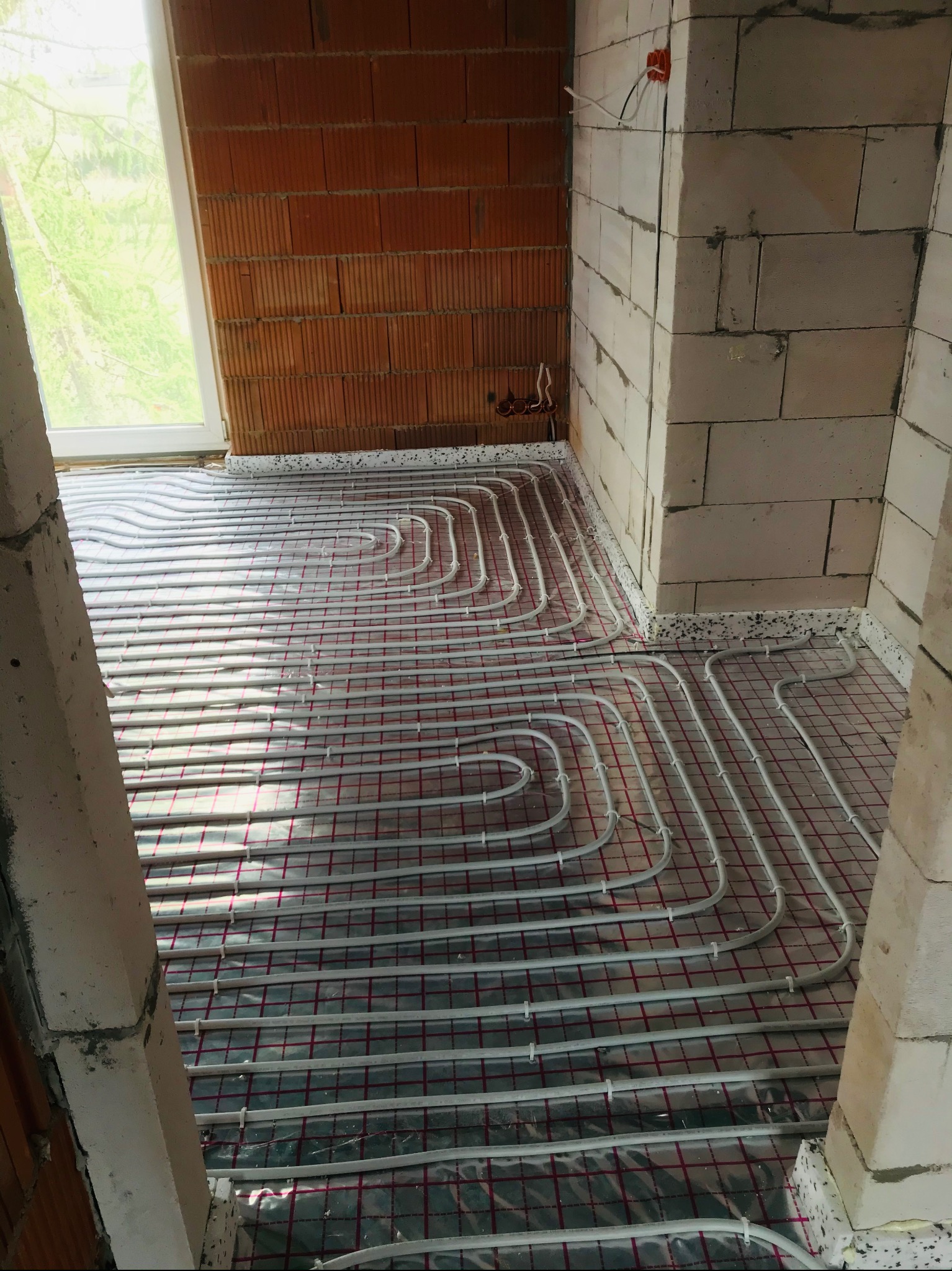 Białe rury ogrzewania podłogowego ułożone na folii z czerwoną kratką w surowym wnętrzu z ceglanymi i betonowymi ścianami w trakcie budowy.