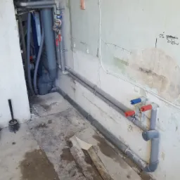 Wnętrze w trakcie remontu z widocznymi szarymi rurami instalacji wodnej i kanalizacyjnej, z zaworami z czerwonymi i niebieskimi uchwytami, przyłącza elektryczne na ścianie i fragmenty starej podłogi.