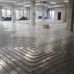 Rozległa powierzchnia z rurami ogrzewania podłogowego ułożonymi na metalowej siatce, widoczne słupy konstrukcyjne budynku i okna w tle, całość przykryta przezroczystą folią ochronną.