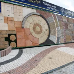 Ekspozycja różnorodnych wzorów i kolorów kostki brukowej, prezentująca aranżacje ścieżek i powierzchni z użyciem elementów betonowych i drewnopodobnych, z logo producenta widocznym na górze.
