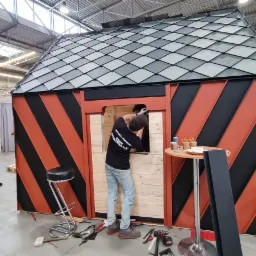 Wnętrze domku pokazowego z ukośnymi pasami w kolorze czarnym i pomarańczowym, pracownik montuje okno, widoczne narzędzia i drabina w tle.