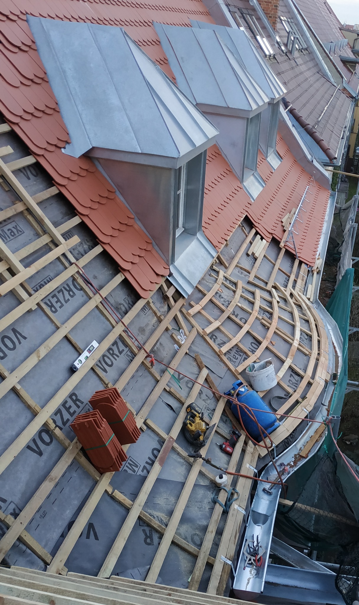 Montaż łat i kontrłat na membranie dachowej, przygotowanie pod dachówkę karpiówkę, widoczne okna dachowe z blachy, narzędzia pracy i fragment rynny.