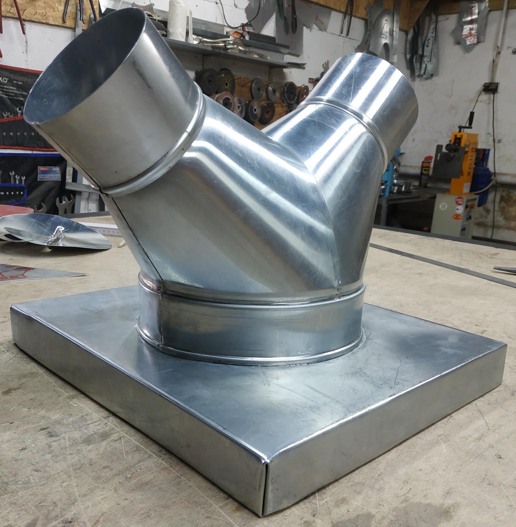 Metalowa kształtka wentylacyjna typu Y na kwadratowej podstawie, wykonana z blachy ocynkowanej, widoczna na warsztatowym stole z narzędziami w tle.