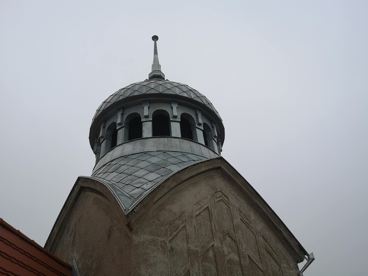 Kopuła z metalową blachą w romby zwieńczona iglicą, widoczna z dołu na tle pochmurnego nieba, z fragmentem ściany budynku o szorstkiej fakturze i czerwonym dachem w rogu kadru.