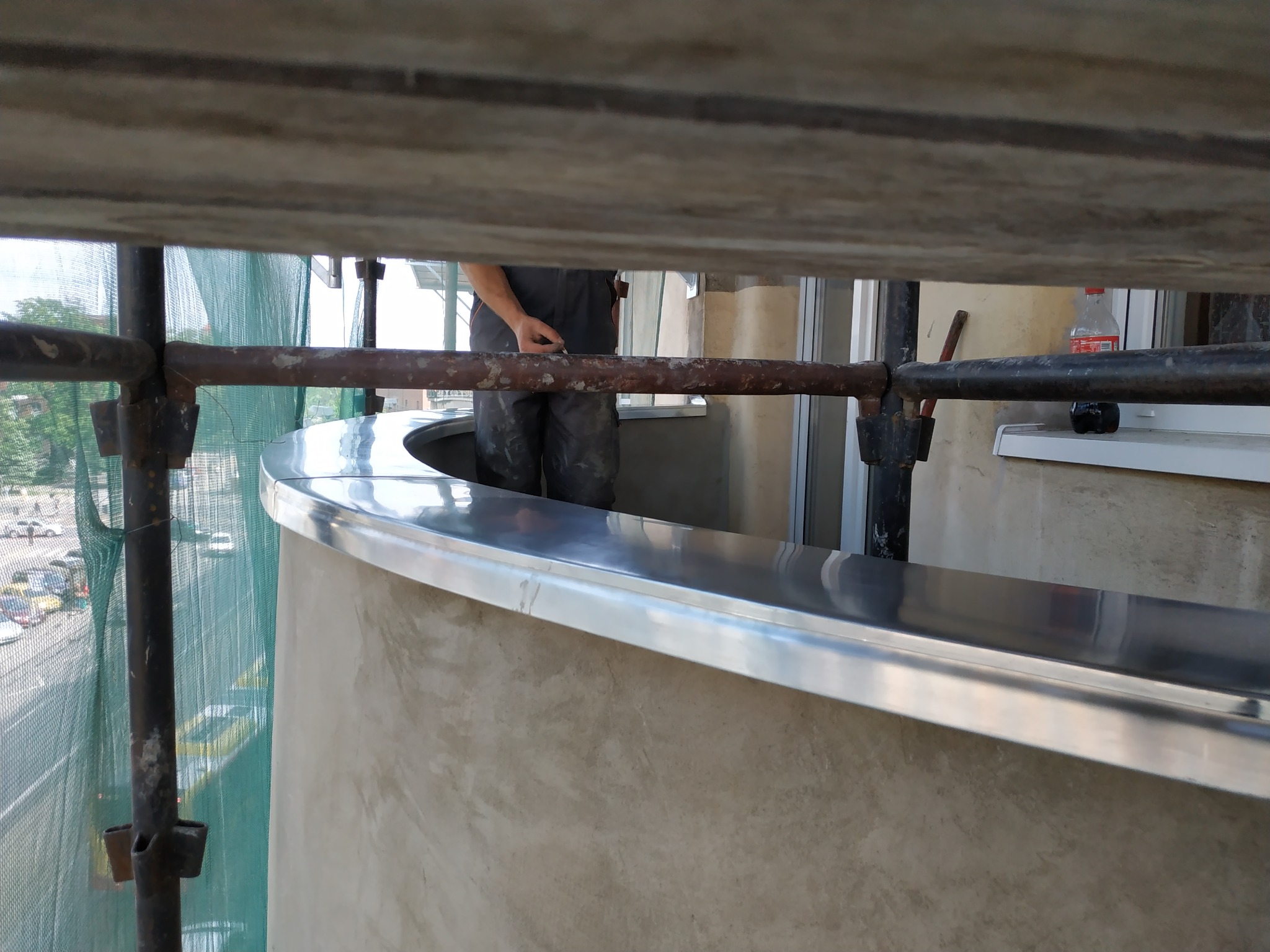 Montaż błyszczącej, metalowej listwy wykończeniowej na balkonie, widok z rusztowania, pracownik w kombinezonie w tle.