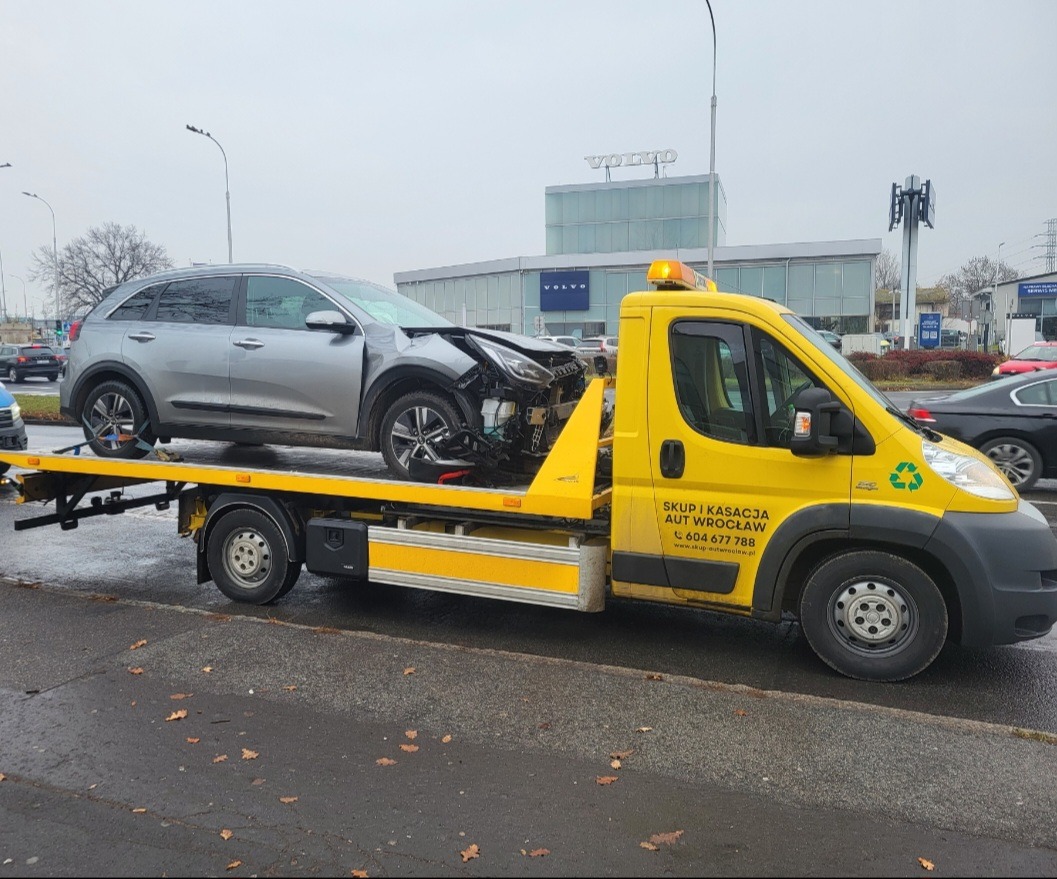 Uszkodzony srebrny SUV na żółtej lawecie z napisem 'Skup i kasacja aut Wrocław' na tle salonu Volvo. Widoczne uszkodzenia przodu pojazdu.