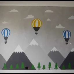 Pokój dziecięcy z ręcznie malowaną ścianą przedstawiającą górski krajobraz z balonami na tle chmur.